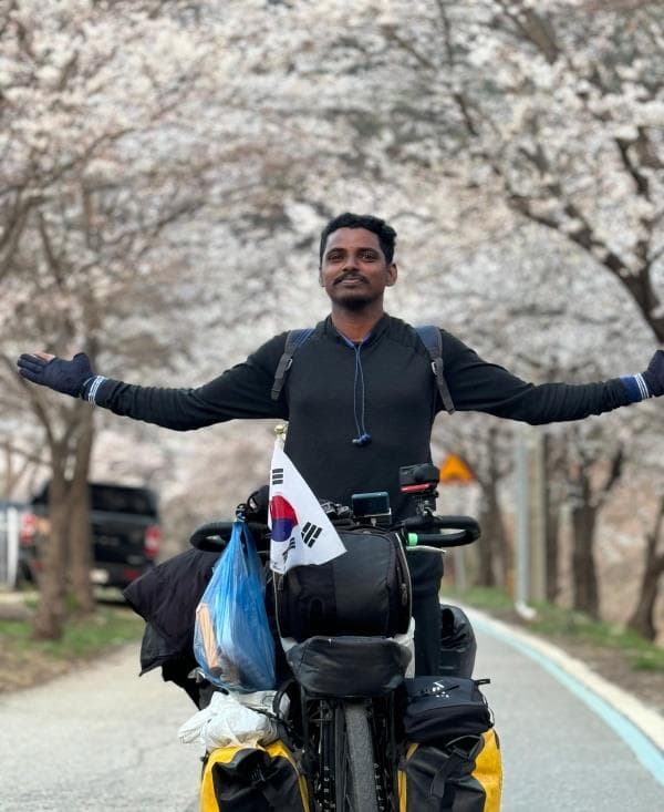 Ranjith standing with his loaded bicycle beneath cherry blossom trees in South Korea
