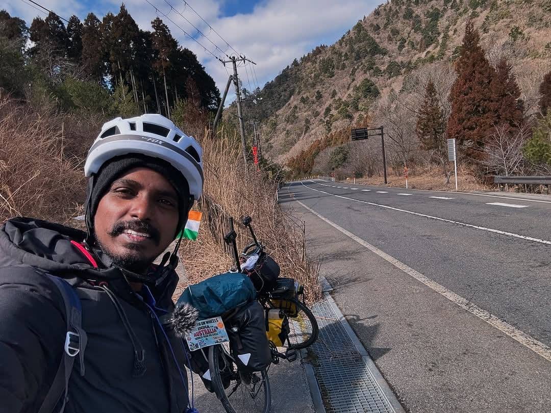 Ranjith cycling a loaded touring bicycle down an open mountain road, Indian flag mounted on the handlebars