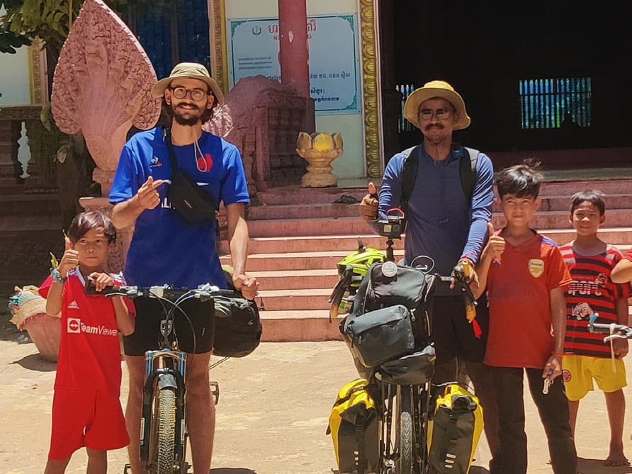Ranjith and a fellow cyclist with their loaded touring bicycles, surrounded by local children, outside a temple in Cambodia