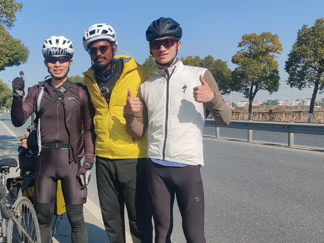 Ranjith and two fellow cyclists in cycling gear and helmets, giving a thumbs up on a tree-lined road in China
