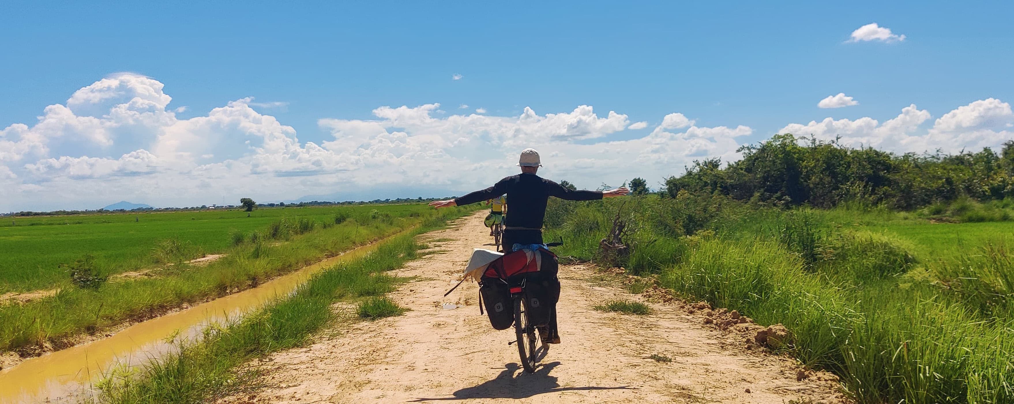 Ranjith riding a loaded touring bicycle with arms outstretched down a farm track in Malaysia
